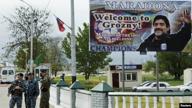 Interior Ministry officers stand beneath a billboard welcoming football legend Diego Maradona to Grozny. Luis Figo, Steve McManaman, and Ivan Zamorano are also scheduled to attend the opening ceremonies.