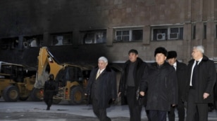 Нұрсұлтан Назарбаев (сол жақтан үшінші) өртеніп кеткен ғимараттың қасында жүр. Жаңаөзен. 22 желтоқсан, 2011 жыл. 