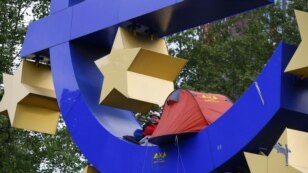 Germaniýaly demonstrant Ýewropanyň Merkezi Bankynyň öňündäki ýewronyň heýkeliniň üstünde oturyp, polisiýanyň protestçileri kowmaga gelerine garaşýar, Frankfurt, 16-njy maý. 