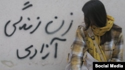 A young Iranian woman sits close to a wall with the slogan "Women, Life, Freedom."
