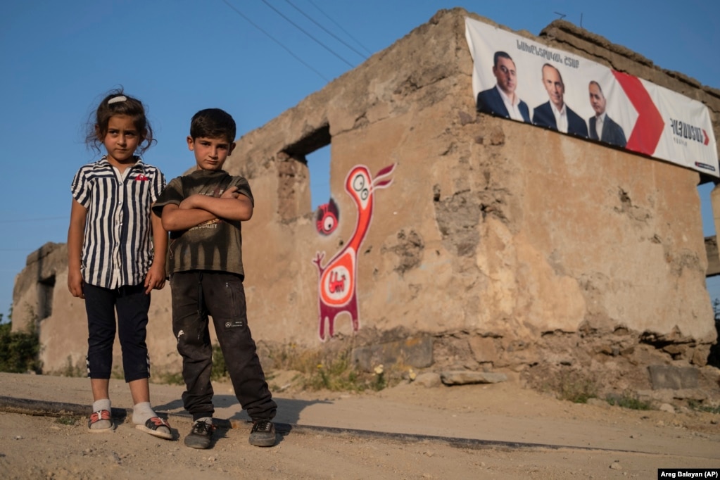 Ерменски деца гледаат во фотограф кој стои пред оштетена зграда со плакати на партиски кандидати во селото Шурнк, на околу 200 километри југоисточно од Ереван, Ерменија, вторник, 15 јуни 2021 година. Селото беше пресечено на два дела од новодефинираната граница со Азербејџан