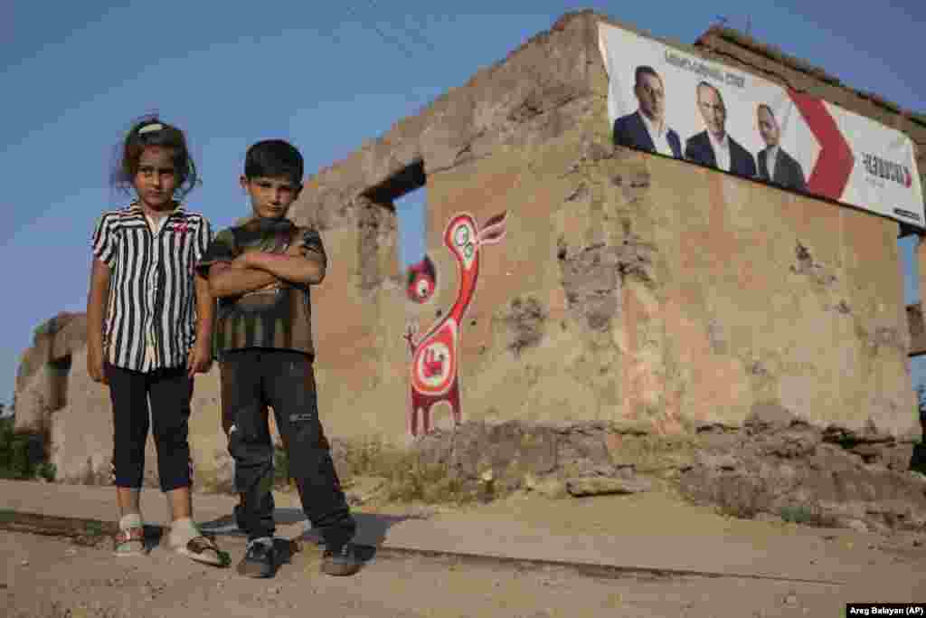Діти у Вірменії дивляться на фотографа, який стоїть перед пошкодженою будівлею з плакатами партійних кандидатів у селі Шурнух, приблизно за 200 км на південний схід від Єревана, Вірменія, 15 червня 2021 року.&nbsp; Село було розділене на дві частини нещодавно визначеним кордоном з Азербайджаном після конфлікту у регіоні Нагірного Карабаху і за мирною угодою втратило цей свій будинок