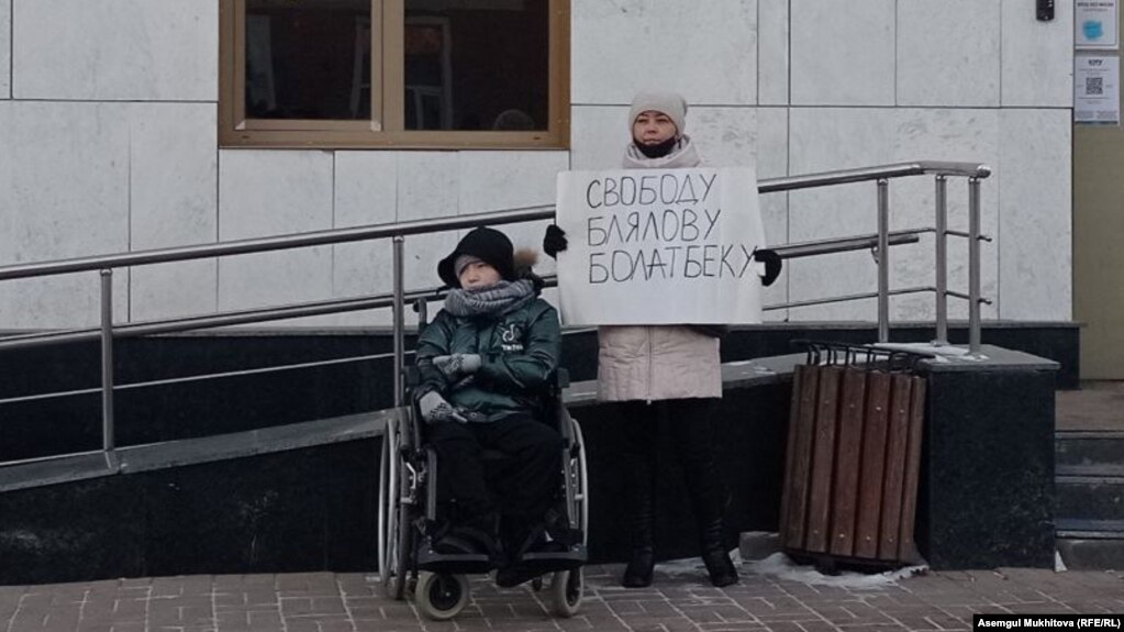 Акмарал Блялова, жена активиста Болатбека Блялова, арестованного на 15 суток, с плакатом стоит вместе с дочерью перед зданием Генеральной прокуратуры. Нур-Султан, 13 декабря 2021 года 