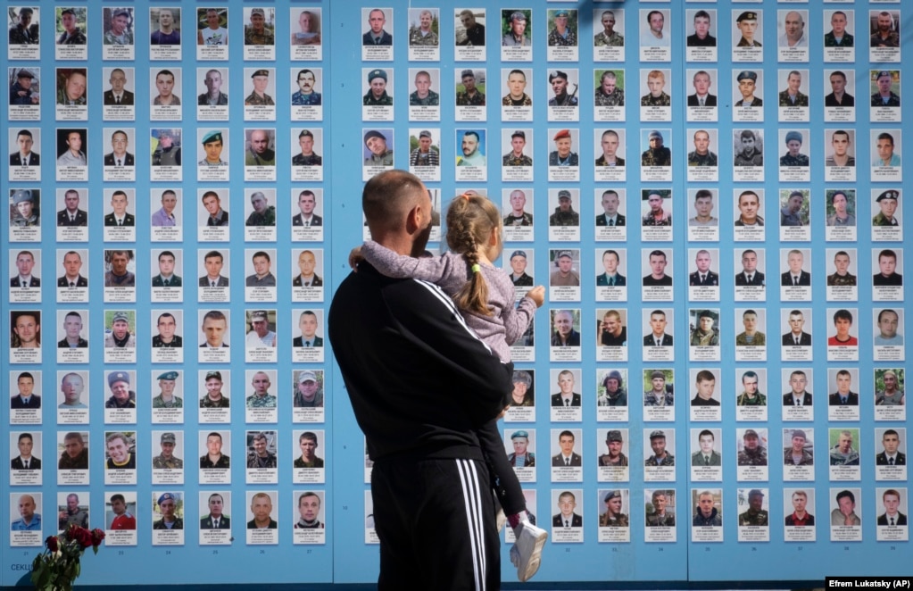 Një burrë në Kiev mban fëmijën e tij para Murit Përkujtimor për Mbrojtësit e rënë të Ukrainës, duke i treguar asaj fotot e shokëve të tij ushtarë që janë vrarë në luftën e vendit të tij kundër forcave pushtuese ruse.