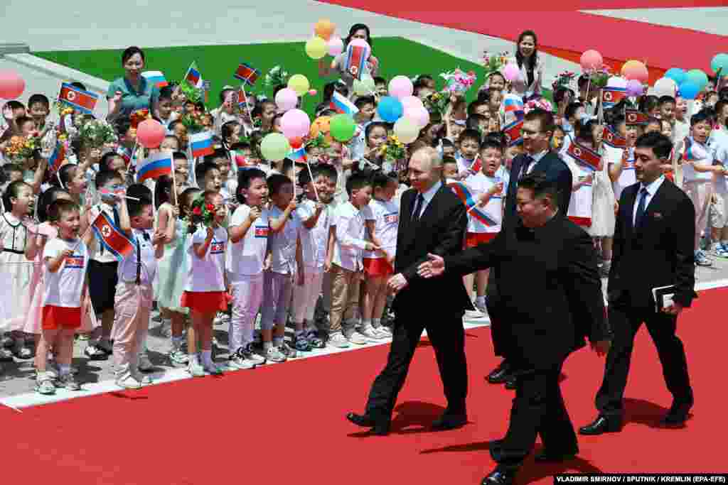 Putin and Kim walk along the red carpet as children greet them waving Russian flags.