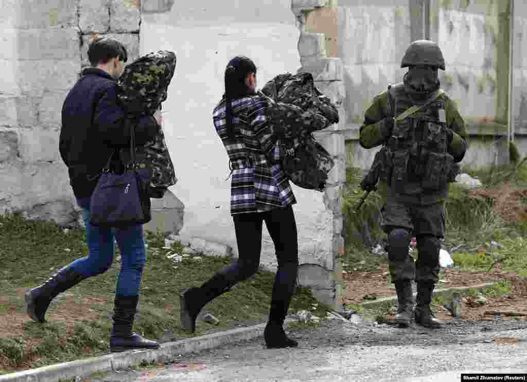 Two women believed to be from the Ukrainian military carry uniforms as they leave a military base and walk past a Russian soldier on March 19. &nbsp;