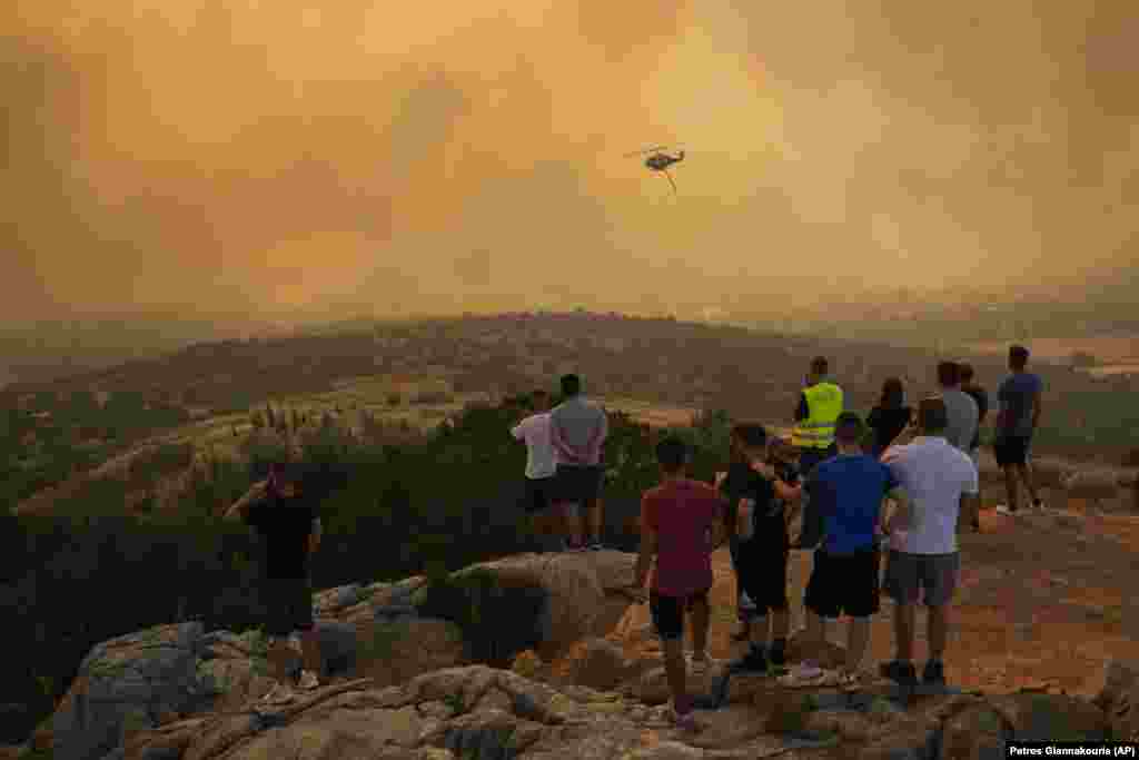 Një helikopter zjarrfikës fluturon nëpër tym ndërsa njerëzit shikojnë në Mandra në perëndim të Athinës, të martën më 18 korrik 2023. Në Greqi, tri vatra të mëdha zjarri dogjën sipërfaqe të tëra jashtë Athinës për të dytën ditë.