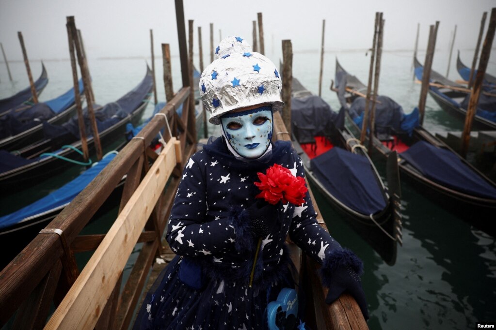 Një pjesëmarrës i veshur me kostum dhe maskë i ulur pranë gondolave në Karnavalin e Venedikut.&nbsp;