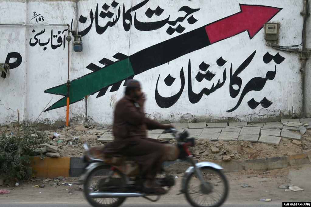 Një motoçiklist kalon pranë një muri ku është pikturuar një shigjetë që është logoja e Partisë Popullore të Pakistanit (PPPP), në një rrugë në Karaçi më 12 janar. Simbolet elektorale vazhdojnë të luajnë një rol të rëndësishëm për t&#39;u siguruar që të dëgjohet zëri i votuesit në procese demokratike.