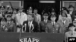 Президентът на САЩ Джон Кенеди изнася реч до американския ген. Лаурис Норщад (вдясно), главнокомандващ на НАТО, на 2 юни 1961 г. в Рокенкур, при посещението във Върховния щаб на съюзническите сили в Европа (SHAPE). Кенеди е убит през 1963 г. в Далас на предизборна обиколка.