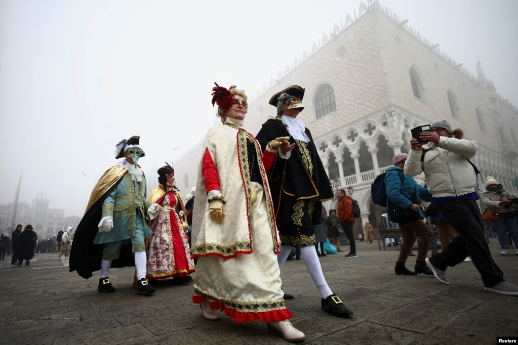 Njerëz të veshur me kostume tradicionale dhe me maska të ndryshme ecin përgjatë Sheshit Shën Marku në Venedik mes turistëve që i shikojnë.