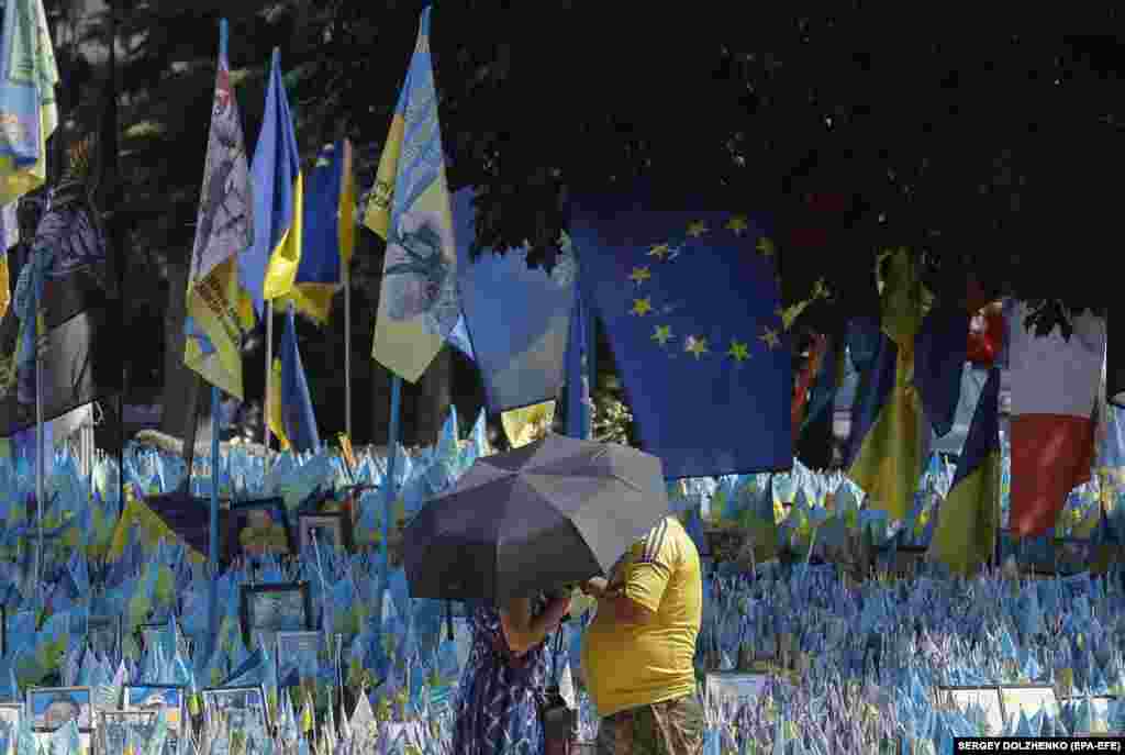 Ukrainians shelter from the sun under an umbrella in front of Ukrainian national flags set up to commemorate fallen soldiers, at Independence Square in Kyiv.