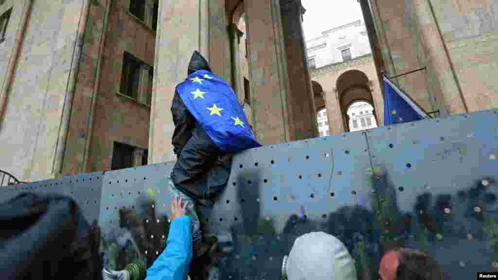 Európai uniós zászlóba burkolózó tüntető a georgiai parlament kerítésén a&nbsp;&bdquo;külföldiügynöktörvény-javaslat&rdquo; elleni tüntetésen Tbilisziben 2024. május 14-én. A kormány egyszer már meghátrált a&nbsp;külföldiügynök-törvény kapcsán, amikor&nbsp;tavaly&nbsp;a tömegtüntetések&nbsp;hatására visszavonta a benyújtott törvényjavaslatot, idén azonban a külföldi ügynök&nbsp;kifejezést kihagyva ismét benyújtotta