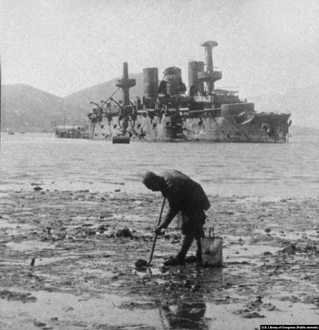 Një burrë duke mbledhur butakë nga balta përpara një anijeje luftarake ruse të shkatërruar në Portin Arthur.