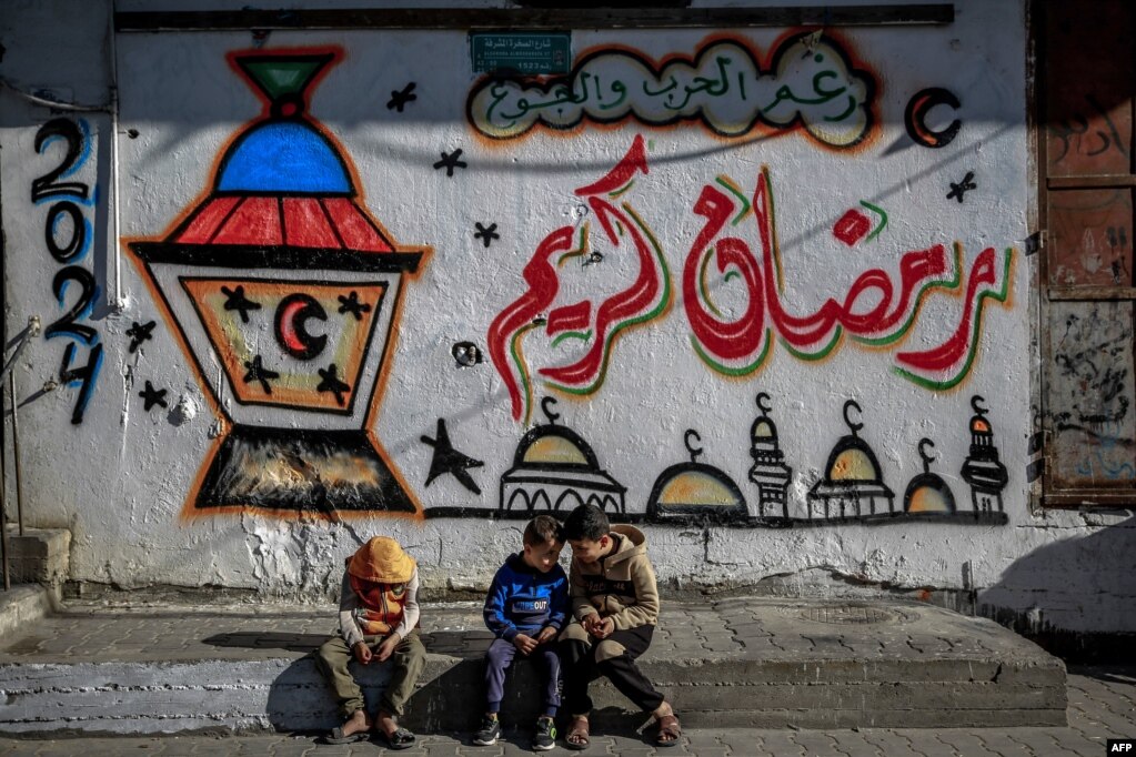 Disa fëmijë palestinezë të ulur pranë një muri në Qytetin e Gazës, ku ka të vizatuara dhe të shkruara mesazhe për Muajin e Ramazanit. 8 mars 2024.&nbsp;