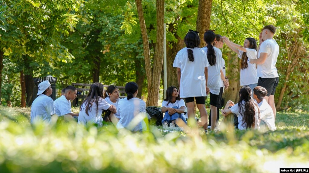 Një grup fëmijësh të ulur shikojnë teksa dy vajza garojnë në një lojë, ku duhet ta gjenin fjalën që kishin të vendosur në ballë në bazë të informatave që merrnin nga garuesja tjetër.