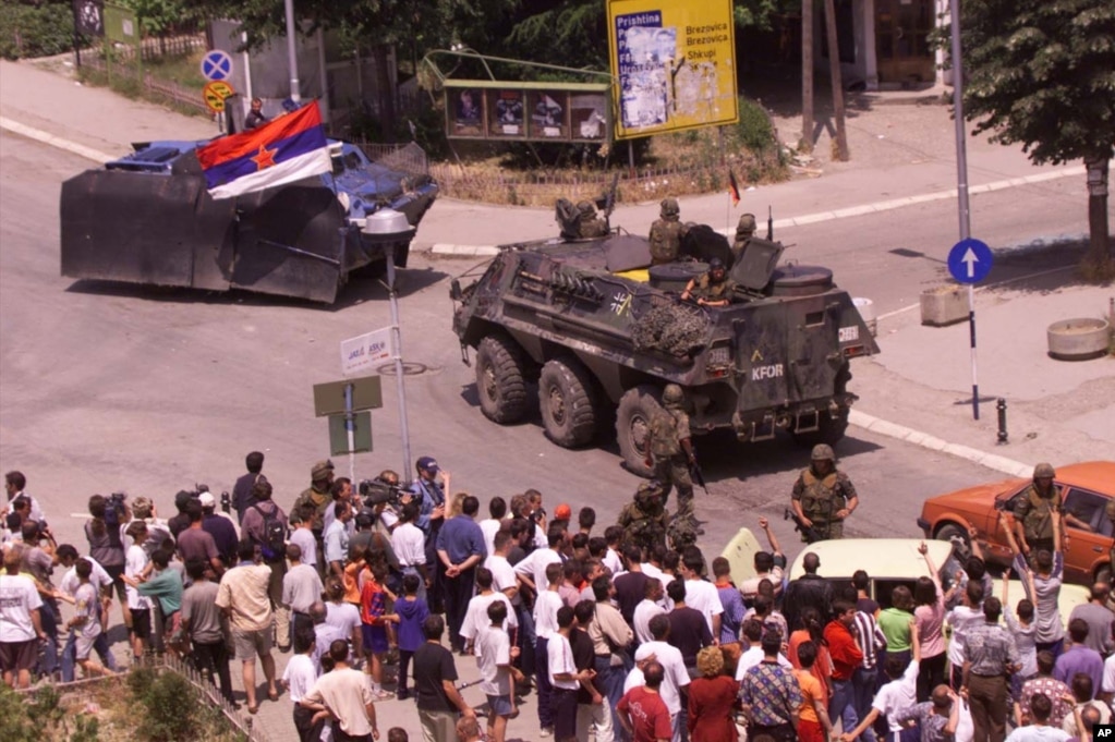 Prizren 14 qershor 1999: Një automjet i modifikuar serb me flamur të Jugosllavisë, shihet derisa largohet nga Prizreni nën mbikëqyrje të forcave paqeruajtëse të KFOR-it gjerman. Shumë qytetarë shihen duke vëzhguar nga afër tërheqjen e trupave serbe. KFOR-i kujdesej për të parandaluar tensionet eventuale dhe përplasjet ndëretnike.   5 qershor 2024: Në udhëkryqin e njëjtë, 25 vjet pas shihet se është larguar tabela rrugore që më 1999 tregonte drejtimet për Prishtinë, Ferizaj, Shkup dhe Brezovicë. 