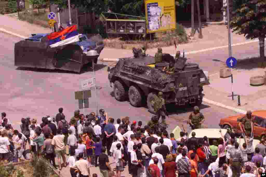 Prizren 14 qershor 1999: Një automjet i modifikuar serb me flamur të Jugosllavisë, shihet derisa largohet nga Prizreni nën mbikëqyrje të forcave paqeruajtëse të KFOR-it gjerman. Shumë qytetarë shihen duke vëzhguar nga afër tërheqjen e trupave serbe. KFOR-i kujdesej për të parandaluar tensionet eventuale dhe përplasjet ndëretnike. &nbsp; 5 qershor 2024: Në udhëkryqin e njëjtë, 25 vjet pas shihet se është larguar tabela rrugore që më 1999 tregonte drejtimet për Prishtinë, Ferizaj, Shkup dhe Brezovicë. 