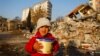 Një fëmijë në Turqi mban në duar një kuti jogurti të marrë nga ndihmat e dërguara, ndërsa në prapavijë shihen ndërtesat e shembura nga tërmeti i 6 shkurtit. 