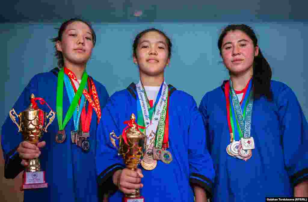 Akibaeva (balra), Bakbur-kizi (középen) és Szofia Zajrova pózol a különböző versenyeken szerzett érmeivel és trófeáival. Zajrova a kirgiz bajnokságon szerzett ezüst- és bronzérmet tartja a kezében, Bakbur-kizi pedig aranyérmet nyert a tizenévesek világbajnokságán &nbsp; 