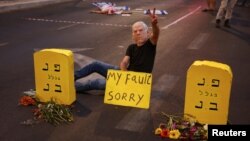  Човек с маска на израелския министър председател Бенямин Нетаняху е лежащ до протестна апаратура от надгробни плочи и държи надпис 