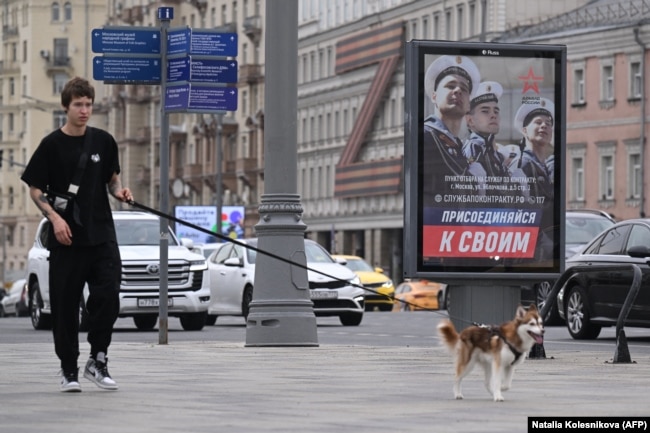 Një djalë i ri duke shëtitur qenin e tij në qendër të Moskës. Afër tij shihet një bilbord i ushtrisë ruse me thirrjen “Bashkojuni njerëzve tuaj”.