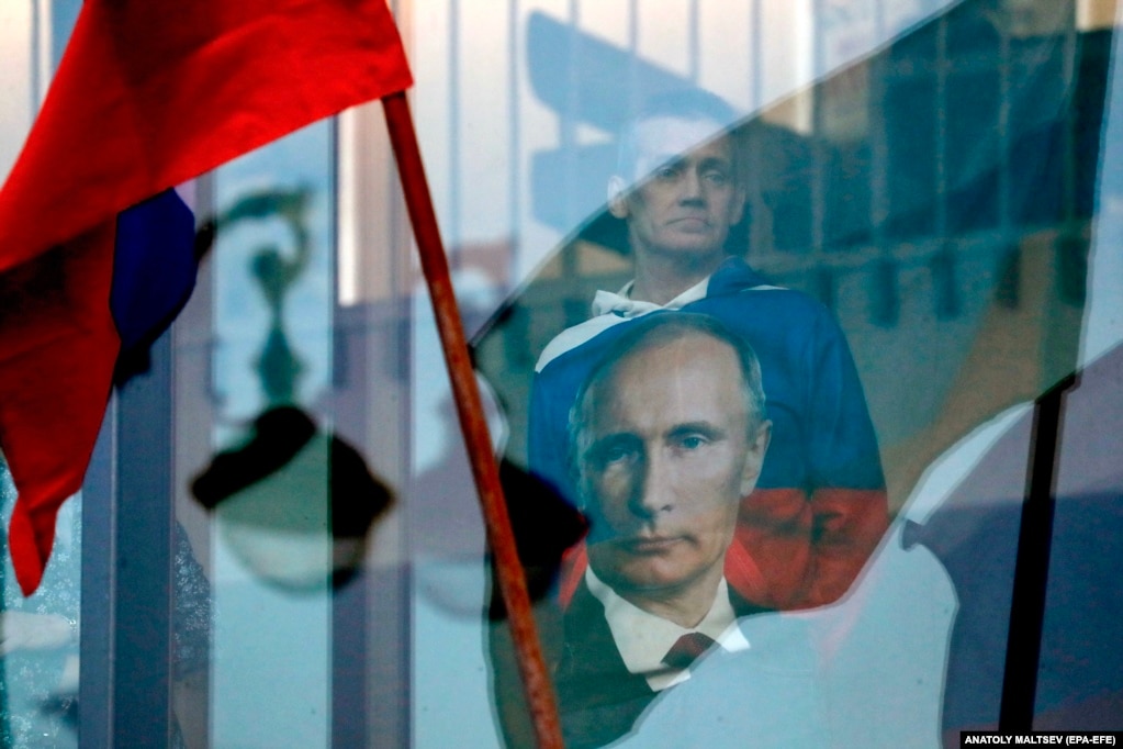 Një person me një bluzë me portretin e presidentit rus, Vladimir Putin, shikon përmes një dritareje, përderisa policia ruan zonën përreth sallës së koncerteve Oktayabrsky në Shën Petersburg.