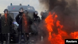 Francia csendőrök és rohamrendőrök sorakoznak fel egy tűz közelében, miközben a tüntetők a Nemzetgyűlés mellett gyülekeznek, hogy tiltakozzanak a nyugdíjreform ellen. Párizs, 2023. március 16.