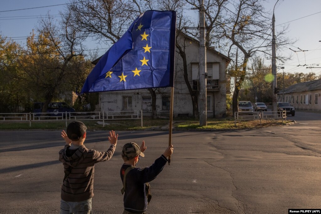 Bambini sventolano una bandiera dell’Unione europea davanti alle auto di passaggio nella città di Kherson, nel sud dell’Ucraina, il 29 ottobre. I residenti nella capitale regionale assediata continuano con le loro vite nonostante i rischi di bombardamenti indiscriminati da parte delle forze di Mosca, che possono mutilare e uccidere in qualsiasi momento.