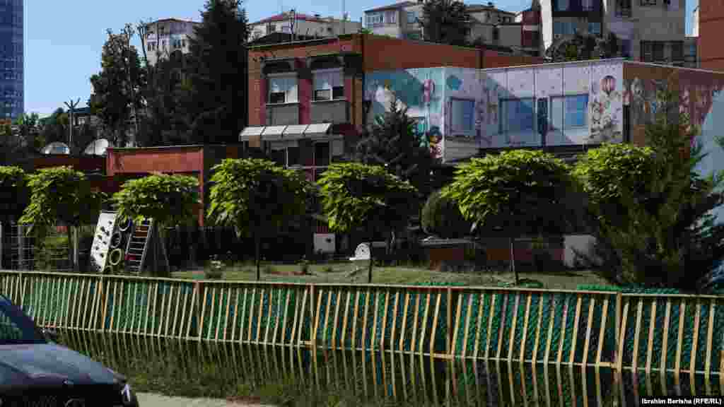 Prishtinë 13 qershor 1999: Ushtarët britanikë derisa shikojnë një objekt banimi kolektiv duke u djegur në Prishtinë. Sipas përshkrimit të fotografisë nga Reuters, kjo fotografi është shkrepur pak kohë pasi trupat britanike ishin sulmuar me armë zjarri nga një pjesëtar i ushtrisë serbe. &nbsp; 5 qershor 2024: Përgjatë viteve shihet se në këtë lokacion është ndërtuar një objekt shkollor, si dhe janë vendosur rrethoja pas zgjerimit të rrugës. Shumë objekte të tjera për banim kolektiv janë ndërtuar në 15 vjetët e fundit. 