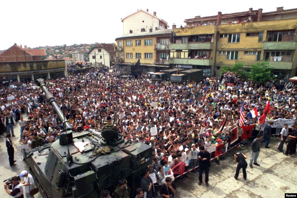 Rahovec 10 korrik 1999:  Në këtë fotografi të publikuar nga Reuters, shihen mijëra qytetarë shqiptarë të Rahovecit derisa protestojnë kundër mundësisë që për sigurinë e tyre të kujdesen trupat ruse që kishin hyrë në Kosovë pa një koordinim me NATO-n.   6 qershor 2024: Sheshi në të cilin u zhvillua protesta, sot ka një pamje krejtësisht tjetër. Një xhami e madhe dhe shumë objekte të reja. 