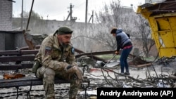 Një ushtar ukrainas shihet i ulur në një stol teksa një banore pastron shkatërrimin që ka afër vetes pas një sulmi ajror rus në rajonin ukrainas Zaporizhja. 5 prill 2024.