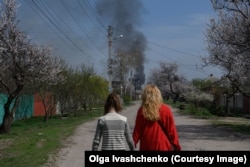 Një grua dhe një vajzë duke ecur nëpër një rrugë në periferi të Harkivit ndërsa para tyre shihet tymi që ngrihet në qiell nga një zjarr, më 9 prill.