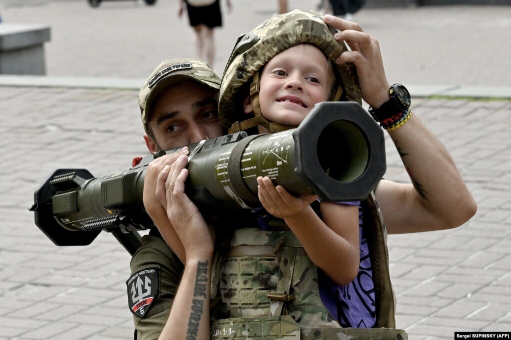 Një ushtar ukrainas ndihmon një djalë të veshur me rroba të blinduara që të mbajë një armë antitank suedeze AT4 në qendër të Kievit më 10 gusht.