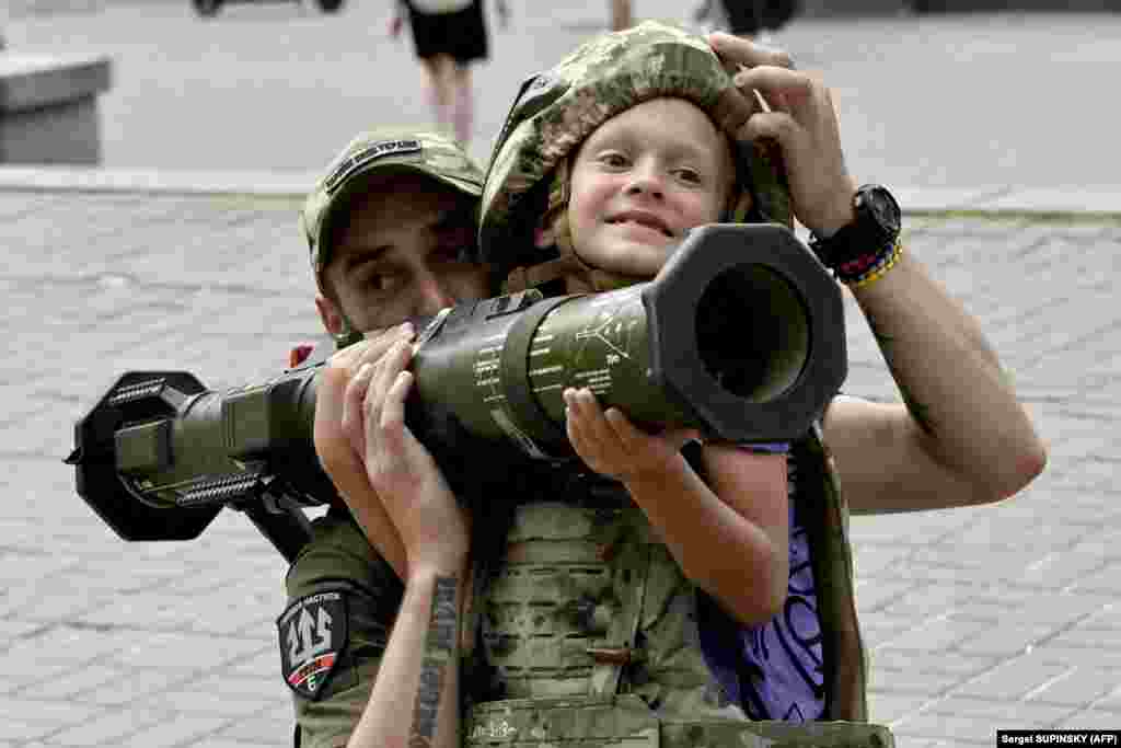 Një ushtar ukrainas ndihmon një djalë të veshur me rroba të blinduara që të mbajë një armë antitank suedeze AT4 në qendër të Kievit më 10 gusht.