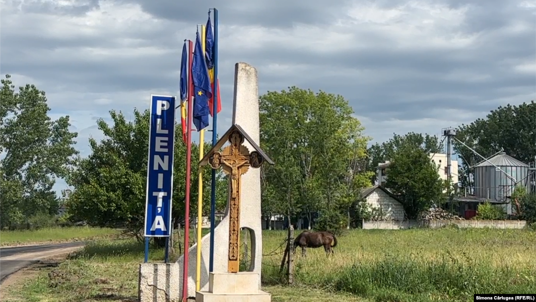 Intrarea în comuna Plenița e simbolică. Are stegurile României, ale Uniunii Europene, o troiță și pe fundal un depozit de cereale.