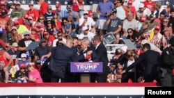  След като се чуват изстрели Тръмп прикляка зад подиума, след което е заобиколен от сътрудници от Сикрет сървис. 