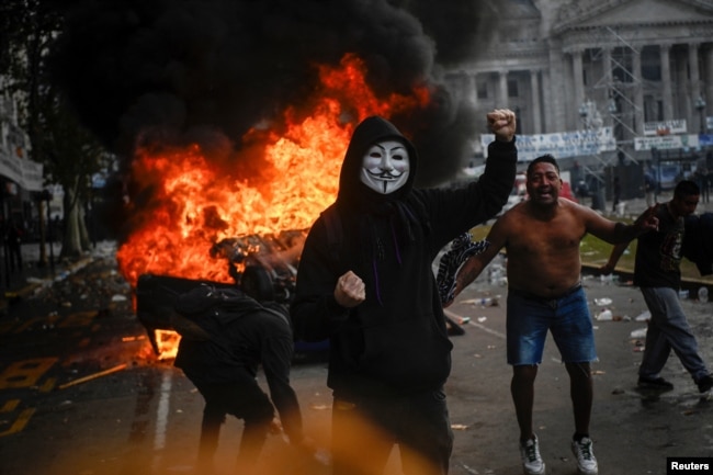 Një veturë duke u djegur teksa një protestues bën gjest me dorë gjatë një proteste kundër projektligjit për reforma në Argjentinë, Buenos Aires, 12 qershor 2024.