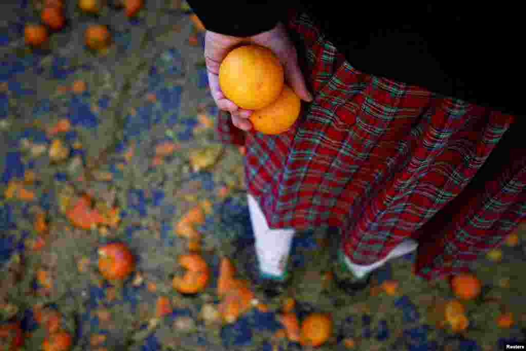 Një pjesëmarrës i veshur me kostum karnavali mban dy portokaj teksa qëndron mes frutave të copëtuara e të shkelura në tokë, Ivrea, Itali, 11 shkurt 2024.