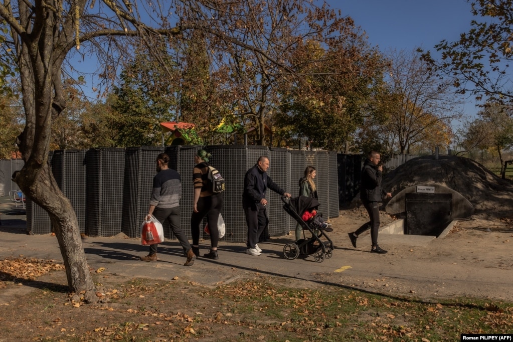 Le persone camminano vicino a un parco giochi per bambini circondato da una recinzione protettiva vicino all'ingresso di un rifugio antiaereo. 