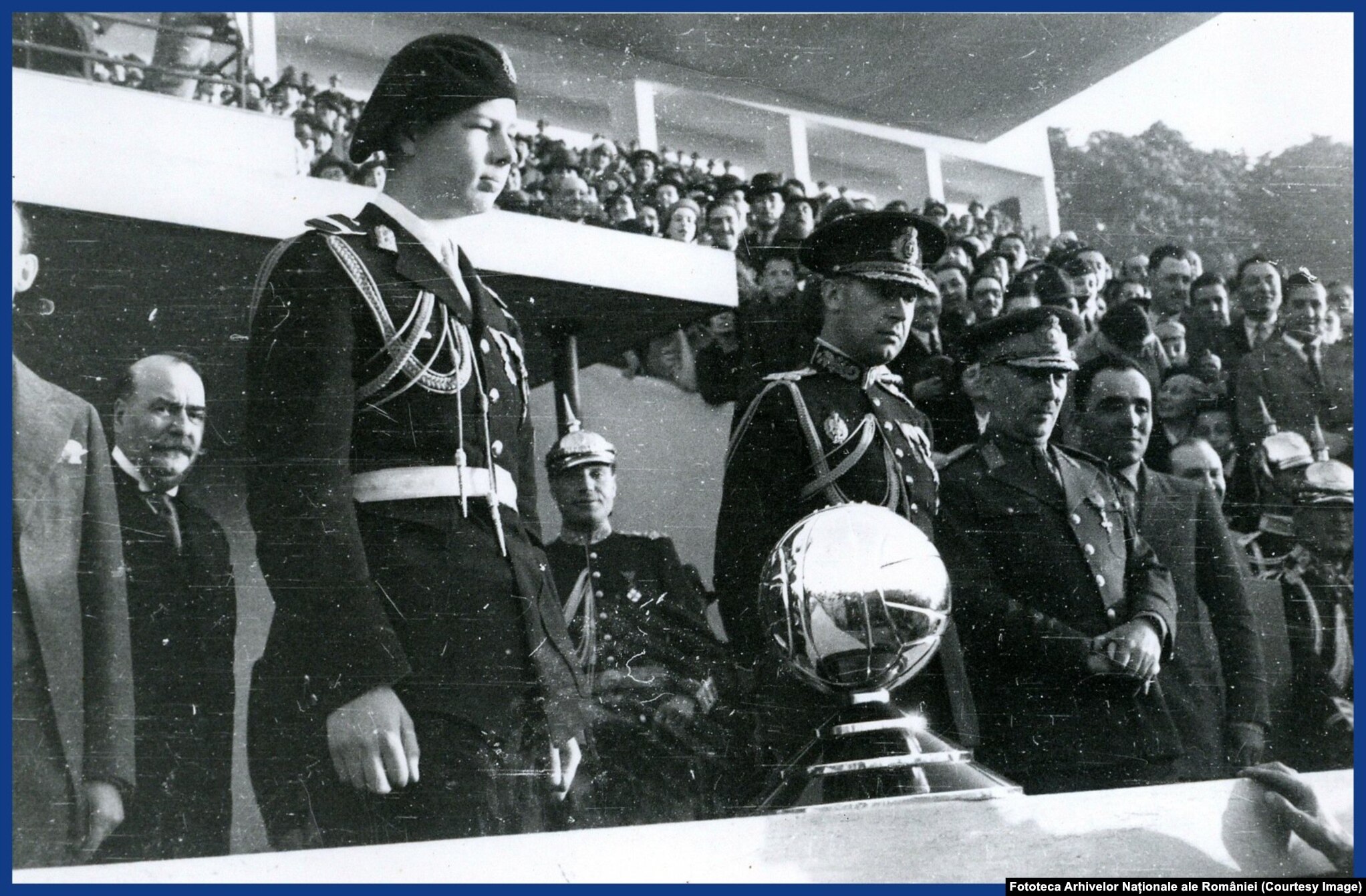 1934. Principele moştenitor Mihai, la un meci de fotbal. În fața sa, un trofeu în forma unei mingi de fotbal.