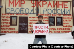Дмитрий Скурихин с плакатом "Прости, Украина" возле своего магазина, фасад которого исписан украинскими городами, пострадавшими из-за военного вторжения. 24 февраля 2023 года