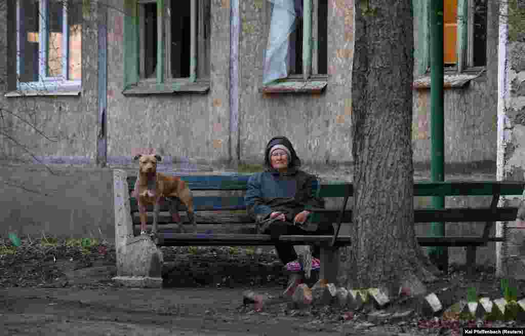 Жінка із собакою сидить на лавці в Часовому Яру. У будинку вибиті усі вікна. 9 квітня 2023 року. Часів Яр, який розташований на височині лише за кілька кілометрів на захід від Бахмута, ймовірно, стане наступним оплотом ЗСУ проти російських сил. Українське командування стверджує, що російські сили у Бахмуті застосовують сирійську тактику&nbsp; &laquo;випаленої землі&raquo;, що знищує все до тла.