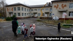 Një mësuese me disa fëmijë ecën përpara një ndërtese të një kopshti, e cila u dëmtua në muajt e parë të sulmit të Rusisë në Ukrainë në qytetin Okhtyrka, rajoni Sumi, Ukrainë, nëntor 2023.