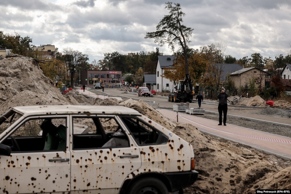 Gli operai di Irpyan stanno ripristinando la strada danneggiata.  Un’auto colpita da colpi di arma da fuoco è un triste ricordo dei feroci combattimenti avvenuti nella periferia di Kiev nel marzo 2022.