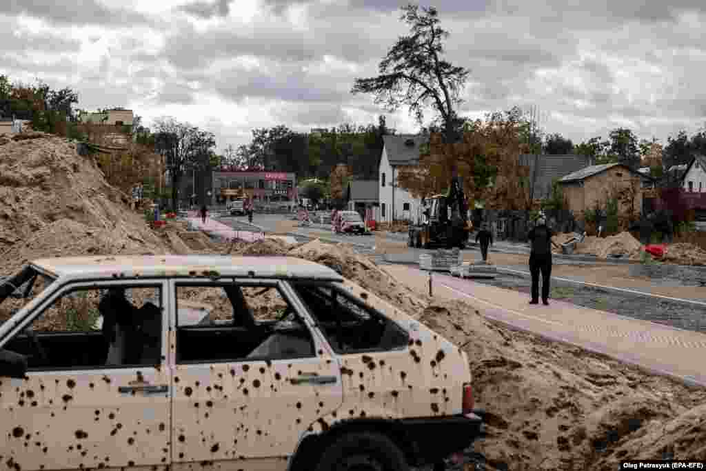 Workers in Irpin&nbsp;rebuild a damaged road on October 23. A shot-up car is a grim reminder of the fierce fighting that took place in the Kyiv suburb in March 2022.