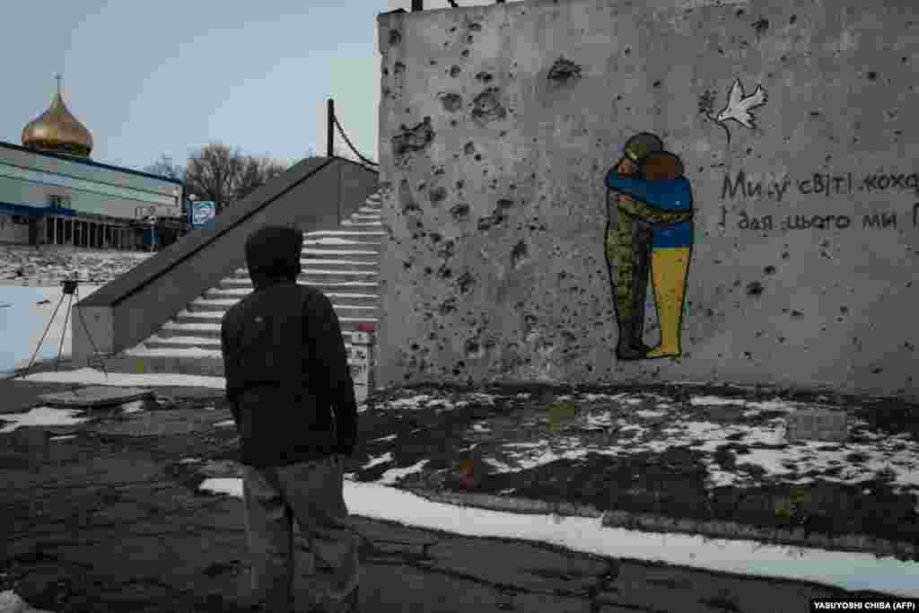 Чоловік дивиться на посічену осколками стіну, на якій намальований мурал з написом: &laquo;Ми у світі кохання помножимо. І для цього ми переможемо!&raquo;. Куп&#39;янськ, Харківська область, 13 лютого 2023 року