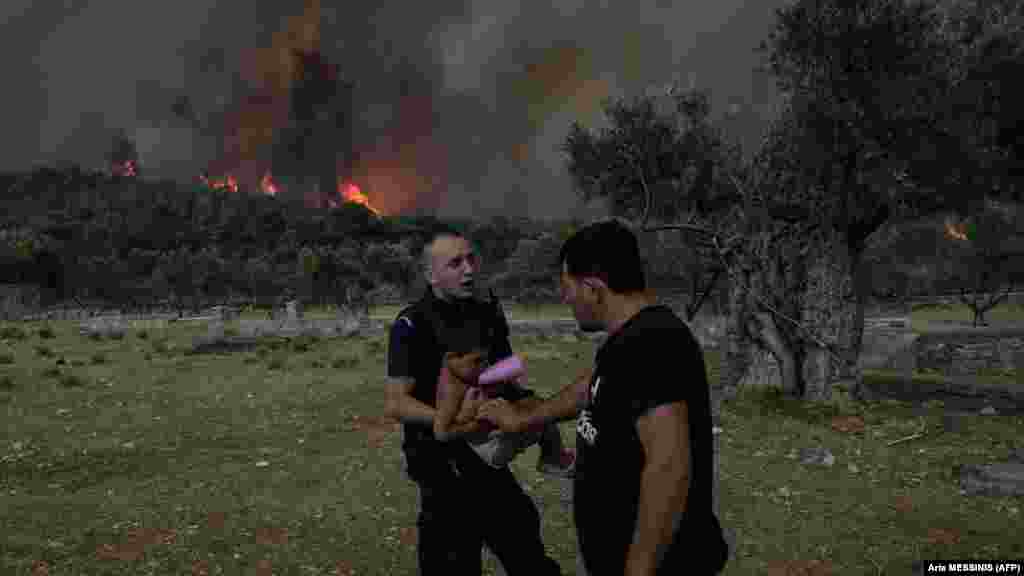 Një oficer policie evakuon një fëmijë ndërsa shumë pranë shihet zjarri që ka përfshirë zonën, fshati Agios Charalampos, pranë Athinës, më 18 korrik 2023.