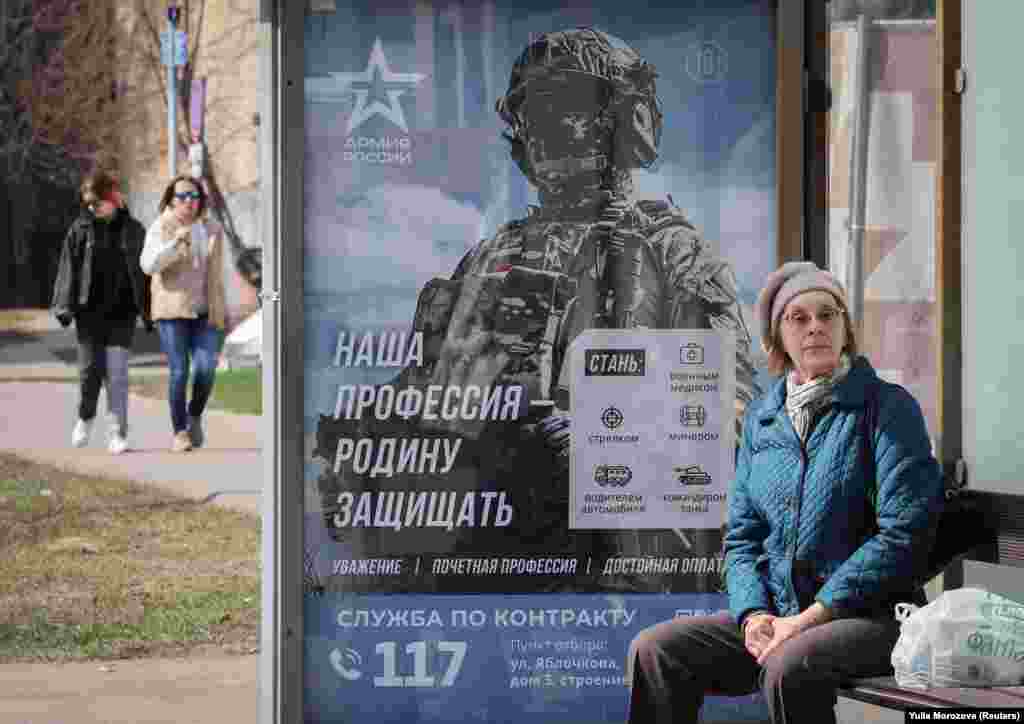 A woman waits at a bus stop next to a poster promoting service in the Russian Army in Moscow.