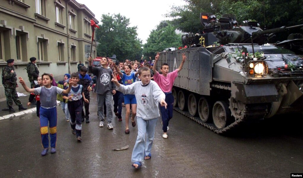 Podujevë 18 qershor 1999: Forcat e KFOR-it arritën në Podujevë disa ditë më vonë se në qytetet tjera të Kosovës. Në këtë fotografi shihen fëmijët duke vrapuar pranë një tanku derisa përshëndesin ushtarët britanikë, të cilët sapo kishin arritur në qytet.   6 qershor 2024: Njëzetepesë vjet pas, në të njëjtin vend objekti komunal mbetet thuajse i njëjtë. Sheshi kryesor është zgjeruar dhe janë ndërtuar objekte të reja. 